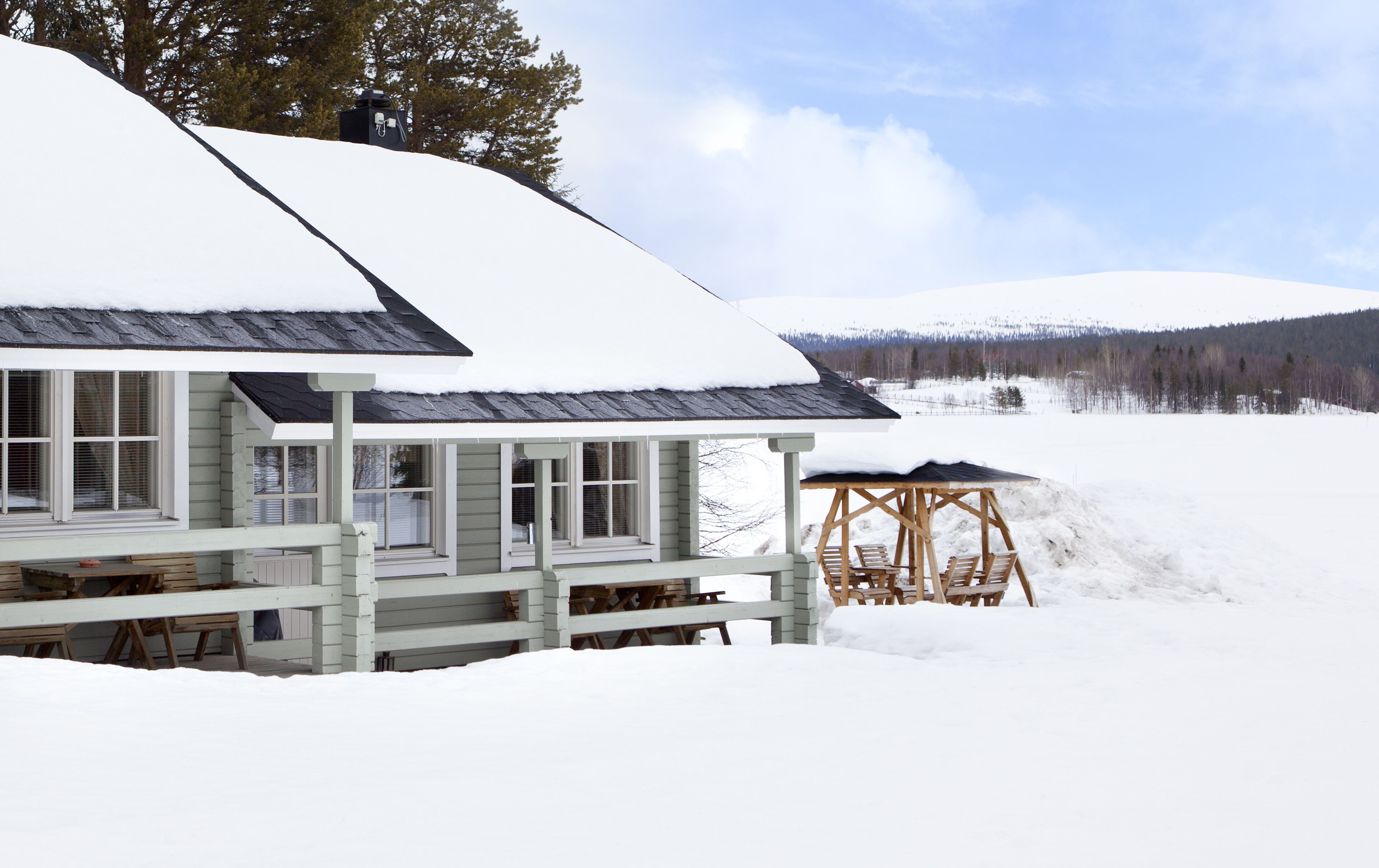 Un chalet dont le toit est recouvert de neige, une terrasse avec des meubles en bois et une vue sur un paysage enneigé, avec des arbres et des collines en arrière-plan.