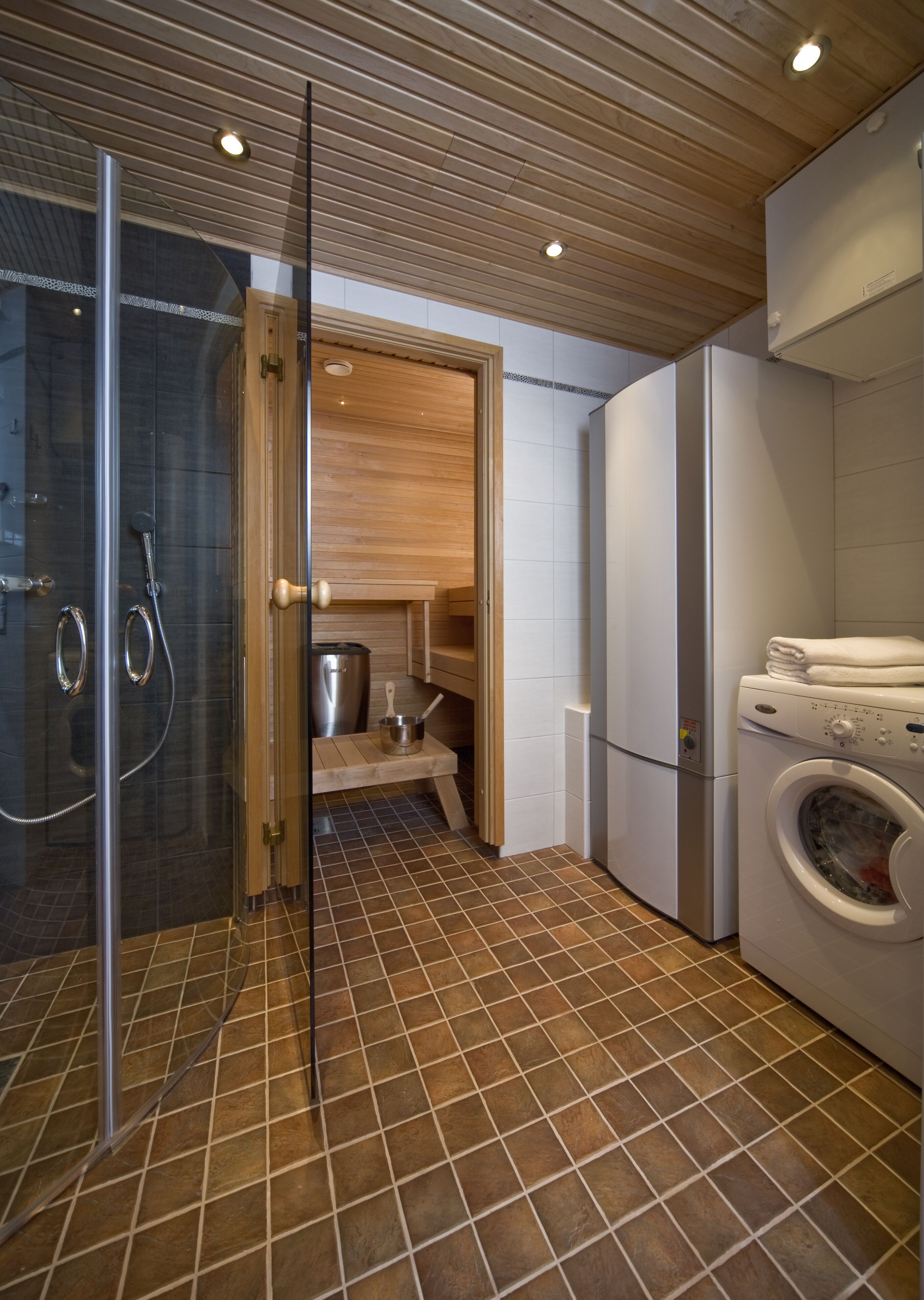 A modern bathroom with a glass shower enclosure, a wooden vanity, a built-in washing machine, and a tiled floor.