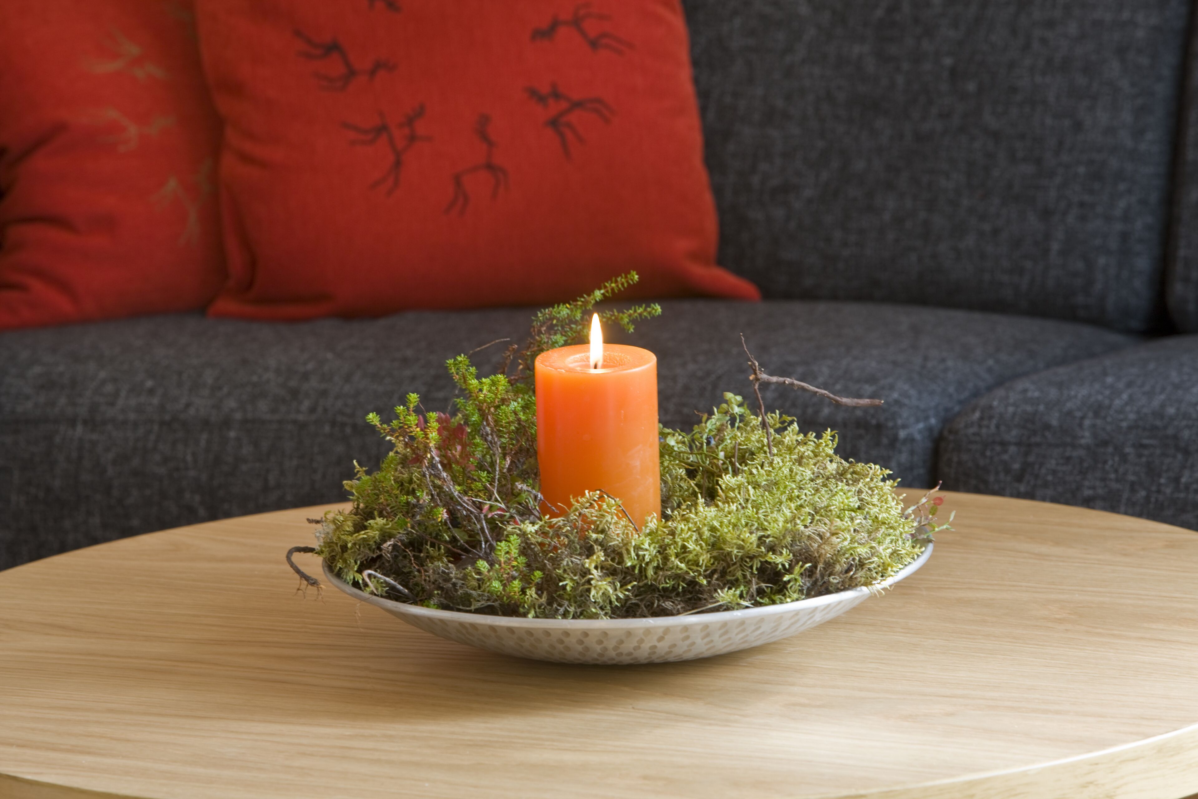 A candle in a bowl of moss on a round table, with a grey sofa and red cushions in the background.