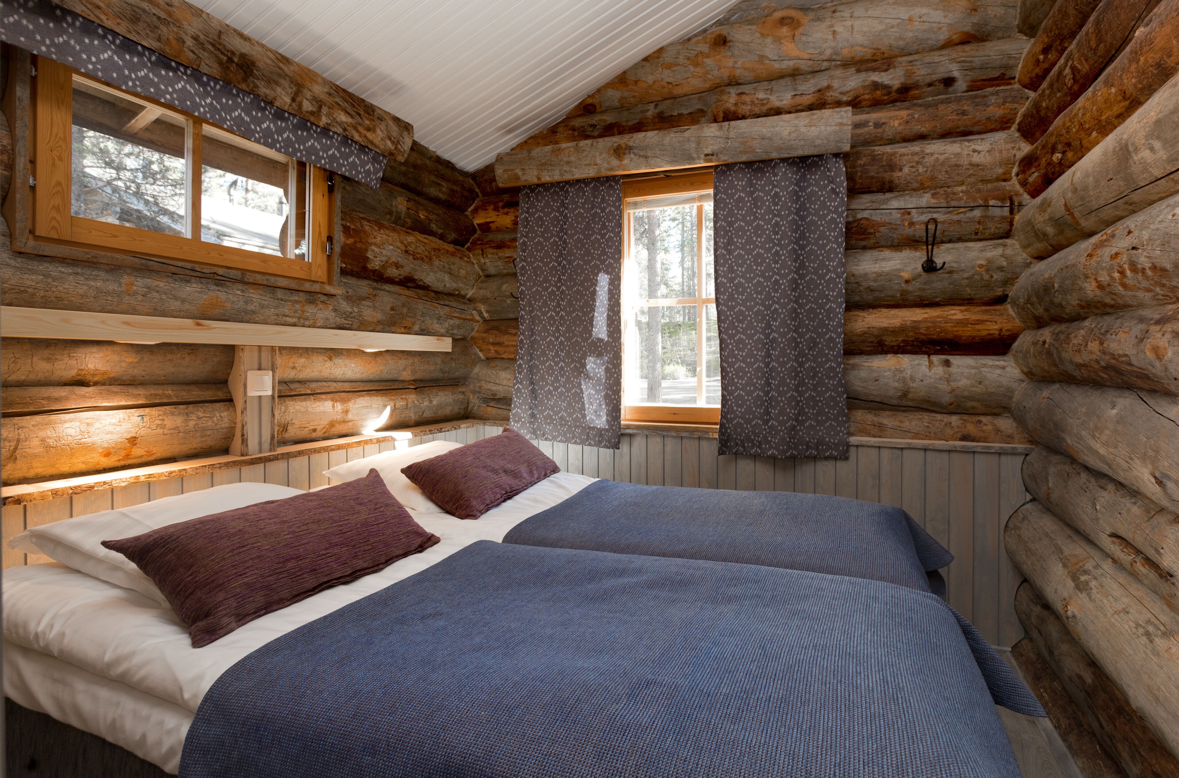 A log cabin room with two beds, a window with curtains, and a wooden headboard.