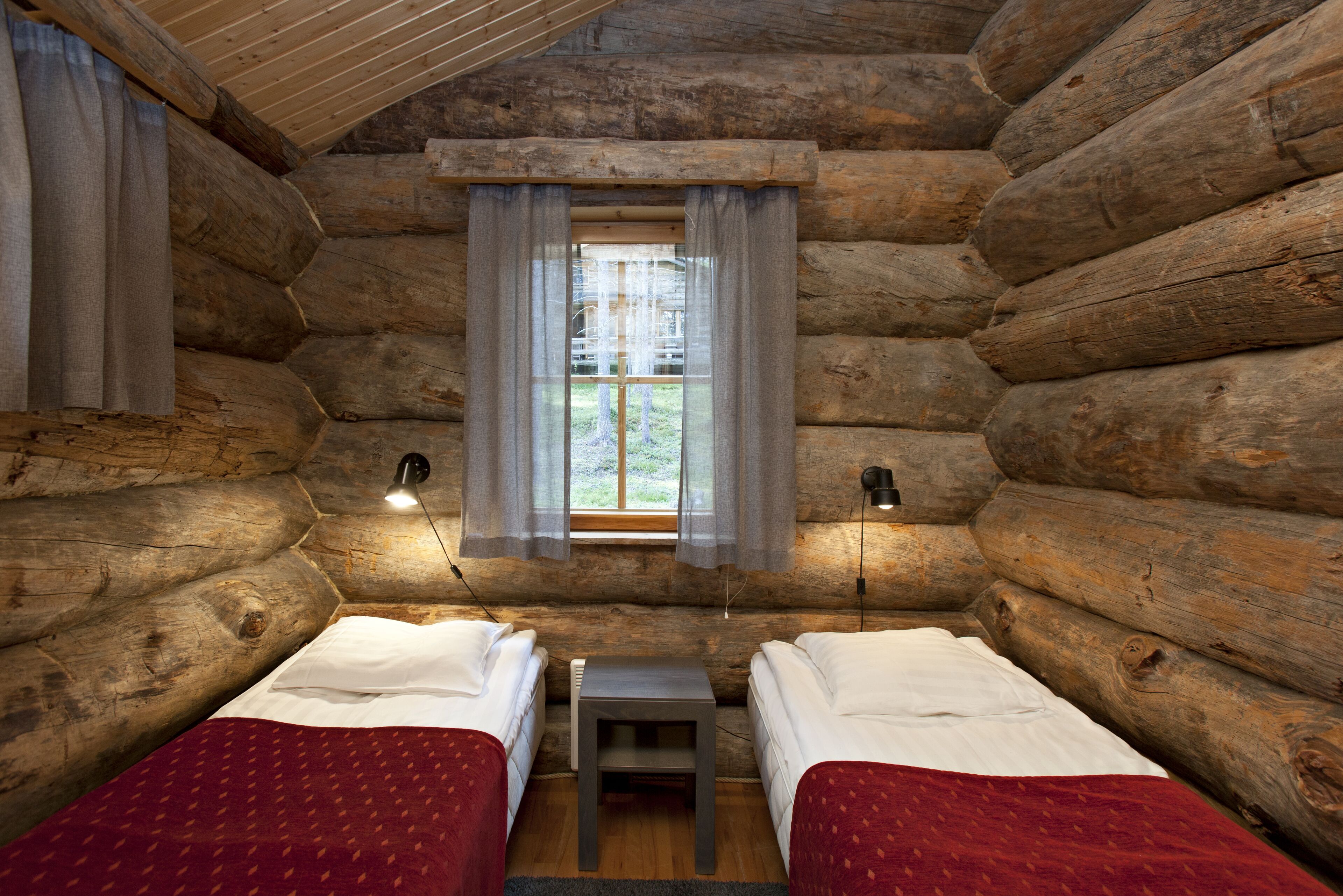 A log cabin room with two beds, a small table, and a window with curtains.