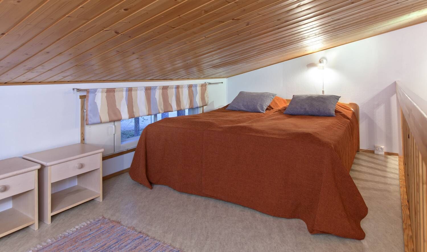 Un dormitorio con techo de madera, una cama con colcha marrón, dos almohadas, una mesita de noche, una lámpara y una ventana con cortina a rayas.