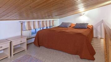 Un dormitorio con techo de madera, una cama con colcha marrón, dos almohadas, una mesita de noche, una lámpara y una ventana con cortina a rayas.