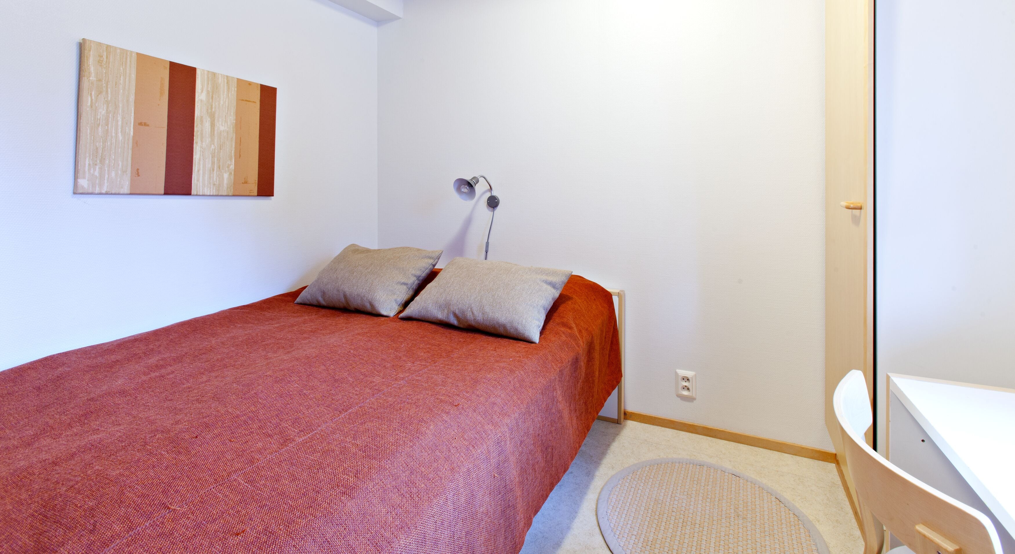 Un dormitorio pequeño y ordenado con una cama individual, dos almohadas, una lámpara de noche, un tapete redondo y un escritorio blanco con una silla.