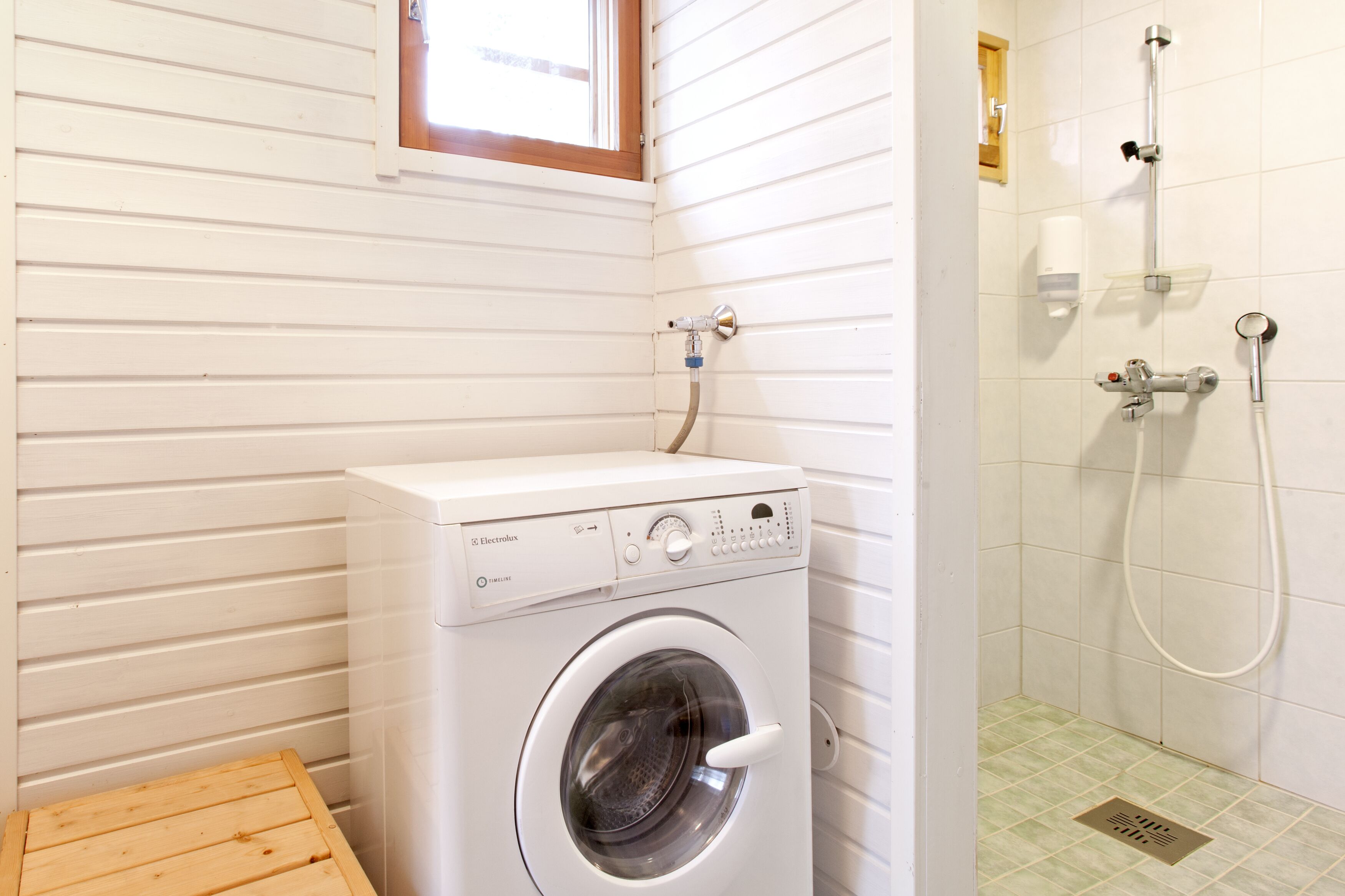 A compact laundry area with a front-loading washing machine, a small wooden stool, and a shower with a handheld hose.