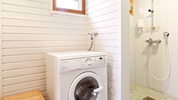 Un espace buanderie compact comprenant un lave-linge à chargement frontal, un petit tabouret en bois et une douche équipée d’un flexible.