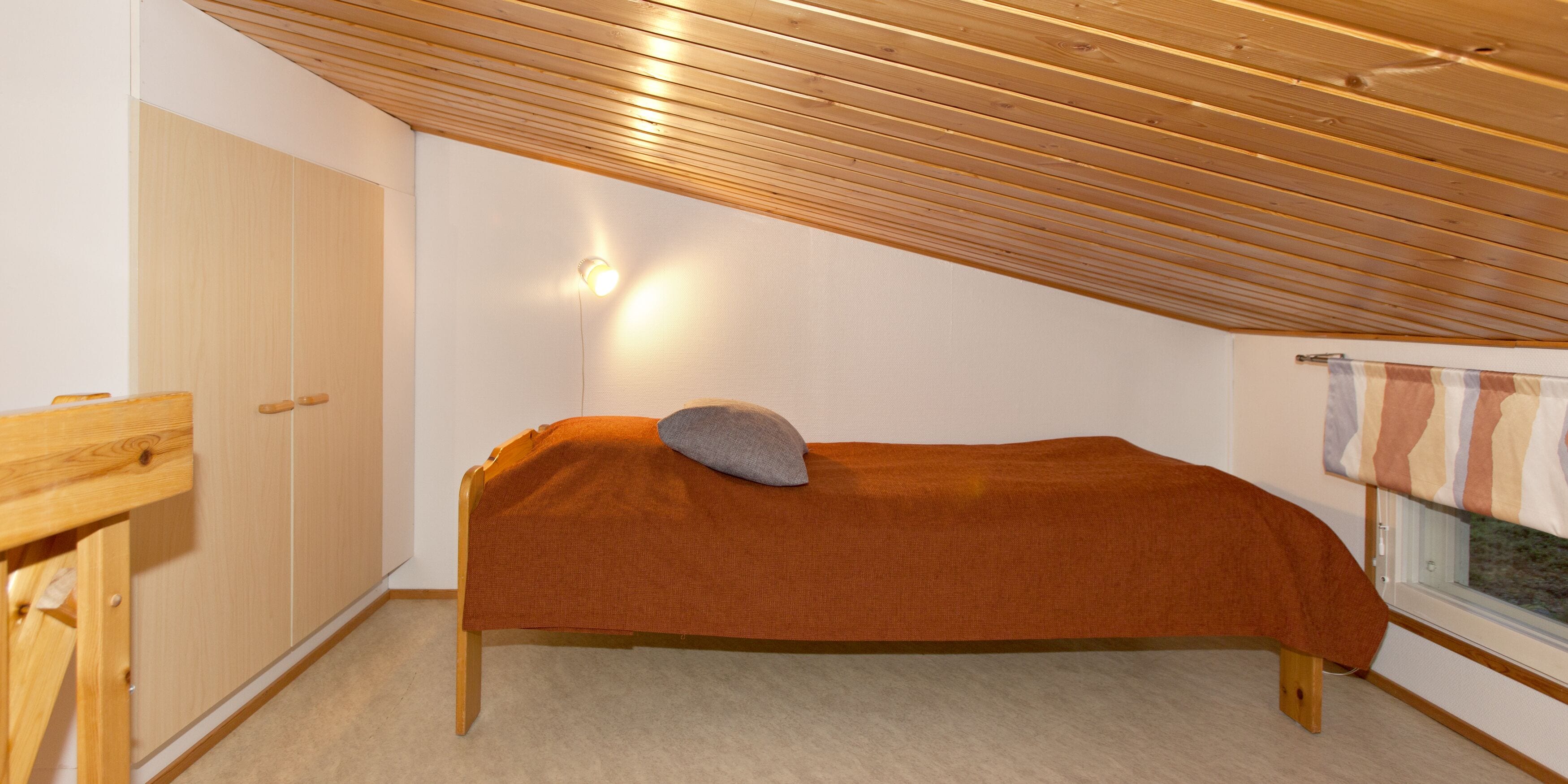 Una habitación pequeña con cama individual, techo inclinado, un armario de madera, una mesita de noche y una ventana con cortina.