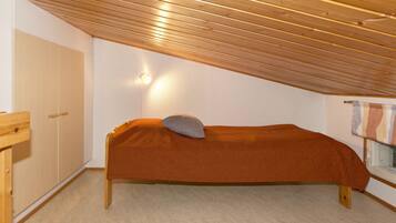 Una habitación pequeña con cama individual, techo inclinado, un armario de madera, una mesita de noche y una ventana con cortina.
