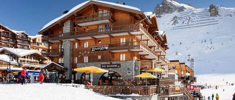 A wooden multi-story building with balconies, a bar/brasserie, and a ski lift in the background.