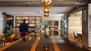 A bar area with a polished wooden counter, shelves with bottles, and a person walking. - Hôtel Le Levanna by Les Etincelles (Tignes)