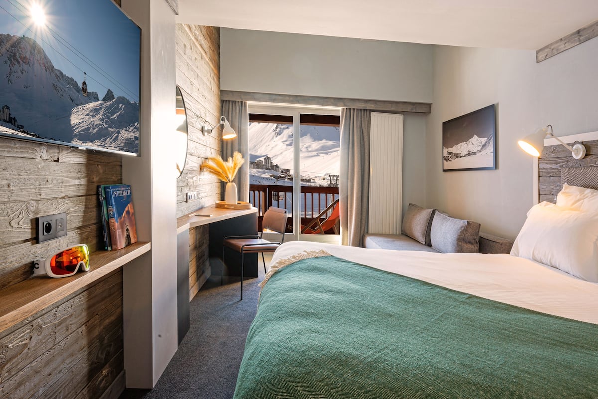 Une chambre d’hôtel avec un lit, un bureau, une chaise, une fenêtre donnant sur les montagnes et, au loin, un télésiège.