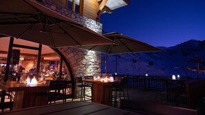 An outdoor dining area with stone walls, wooden tables, and chairs, overlooking a snowy mountain. - Hôtel Le Levanna by Les Etincelles (Tignes)