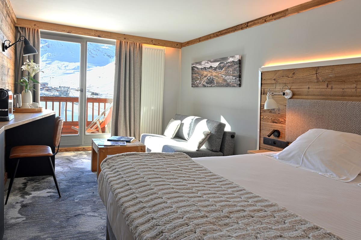 Une chambre d’hôtel avec un lit, un bureau, une chaise, un canapé et une vue sur des montagnes enneigées, visible par une porte coulissante.
