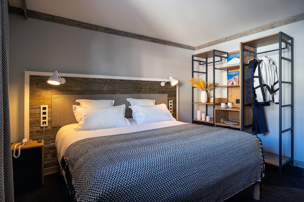 Une chambre d’hôtel bien aménagée, avec un grand lit, des lampes de chevet, une table de chevet, une armoire et une étagère contenant des livres et un manteau.