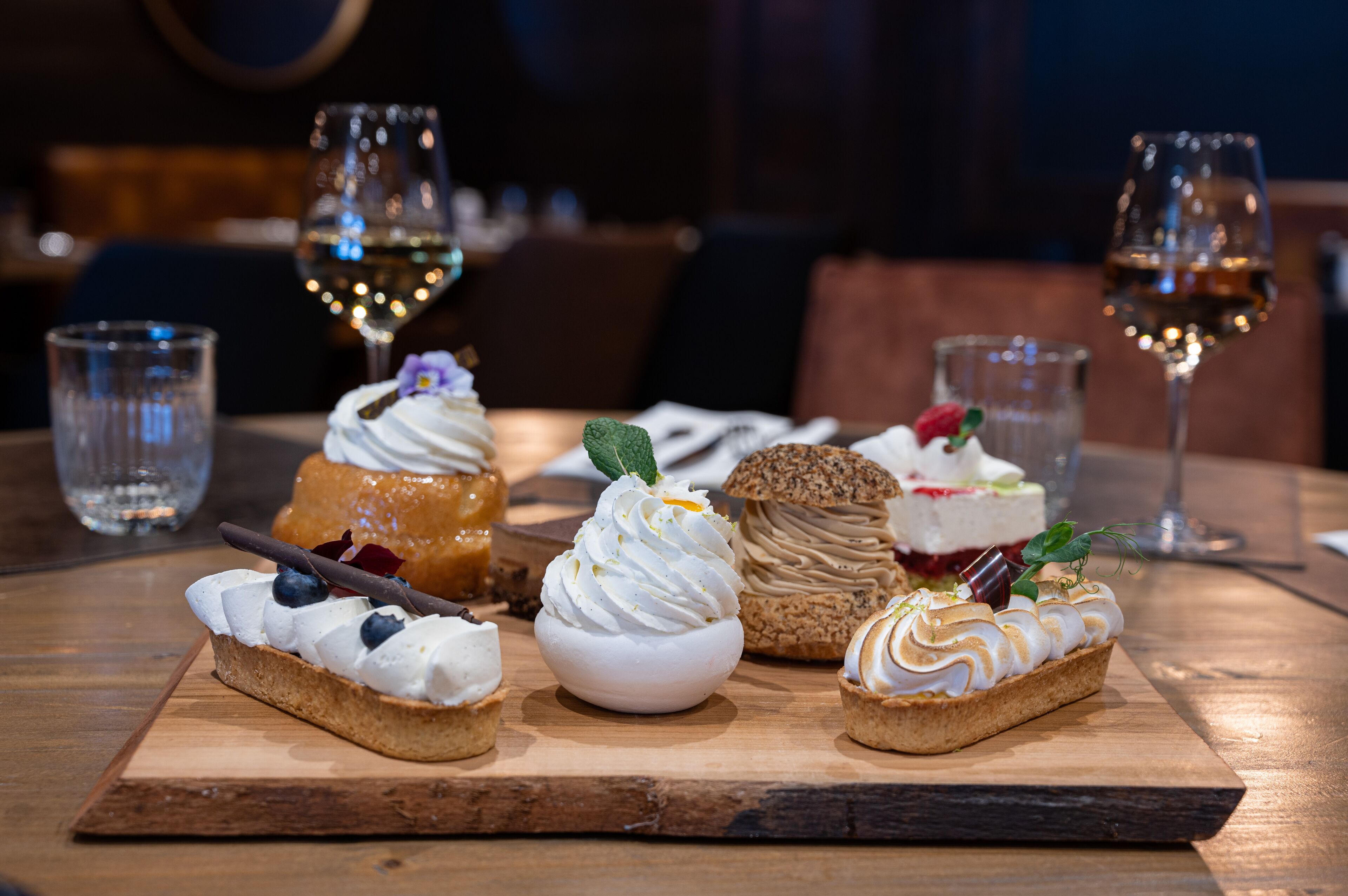 A wooden board with various desserts and glasses of wine on a table.