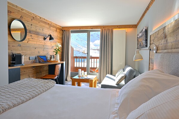 Habitación de hotel con pared de madera, una cama, un escritorio con una computadora, una silla, una mesita con un florero y vistas a una montaña nevada.