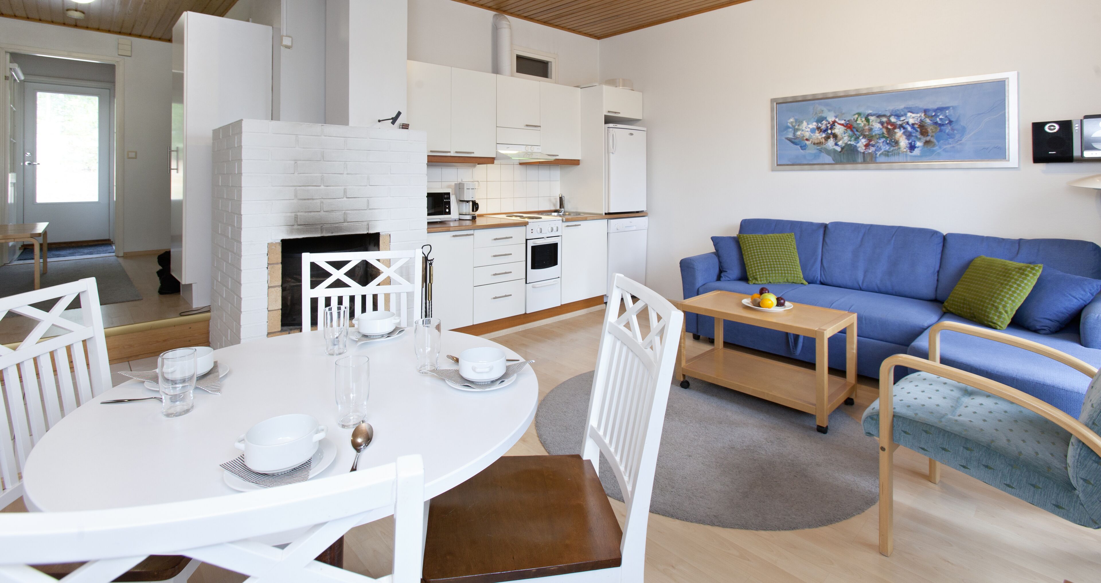 A modern kitchen with white cabinets, a built-in oven, and a white brick fireplace. There is a blue sofa with green cushions and a small wooden table with fruit.