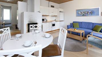 A modern kitchen with white cabinets, a built-in oven, and a white brick fireplace. There is a blue sofa with green cushions and a small wooden table with fruit.