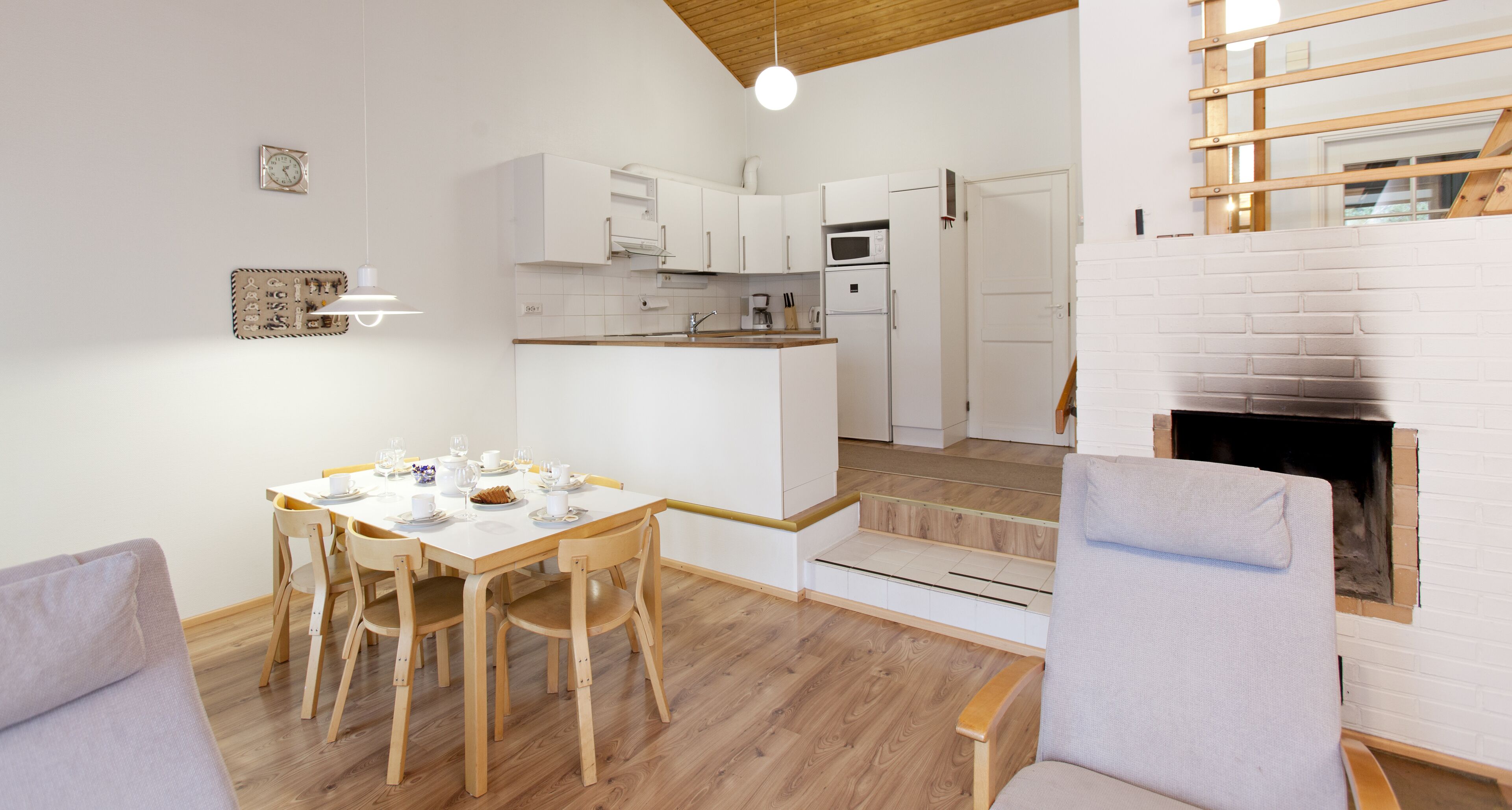 A modern kitchen with white cabinets, a microwave, and a built-in oven. There is a dining area with a wooden table and chairs, and a fireplace with a brick surround.