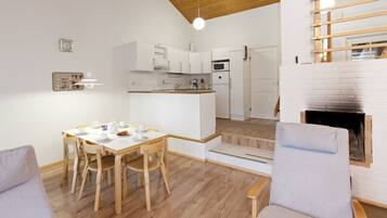 A modern kitchen with white cabinets, a microwave, and a built-in oven. There is a dining area with a wooden table and chairs, and a fireplace with a brick surround.