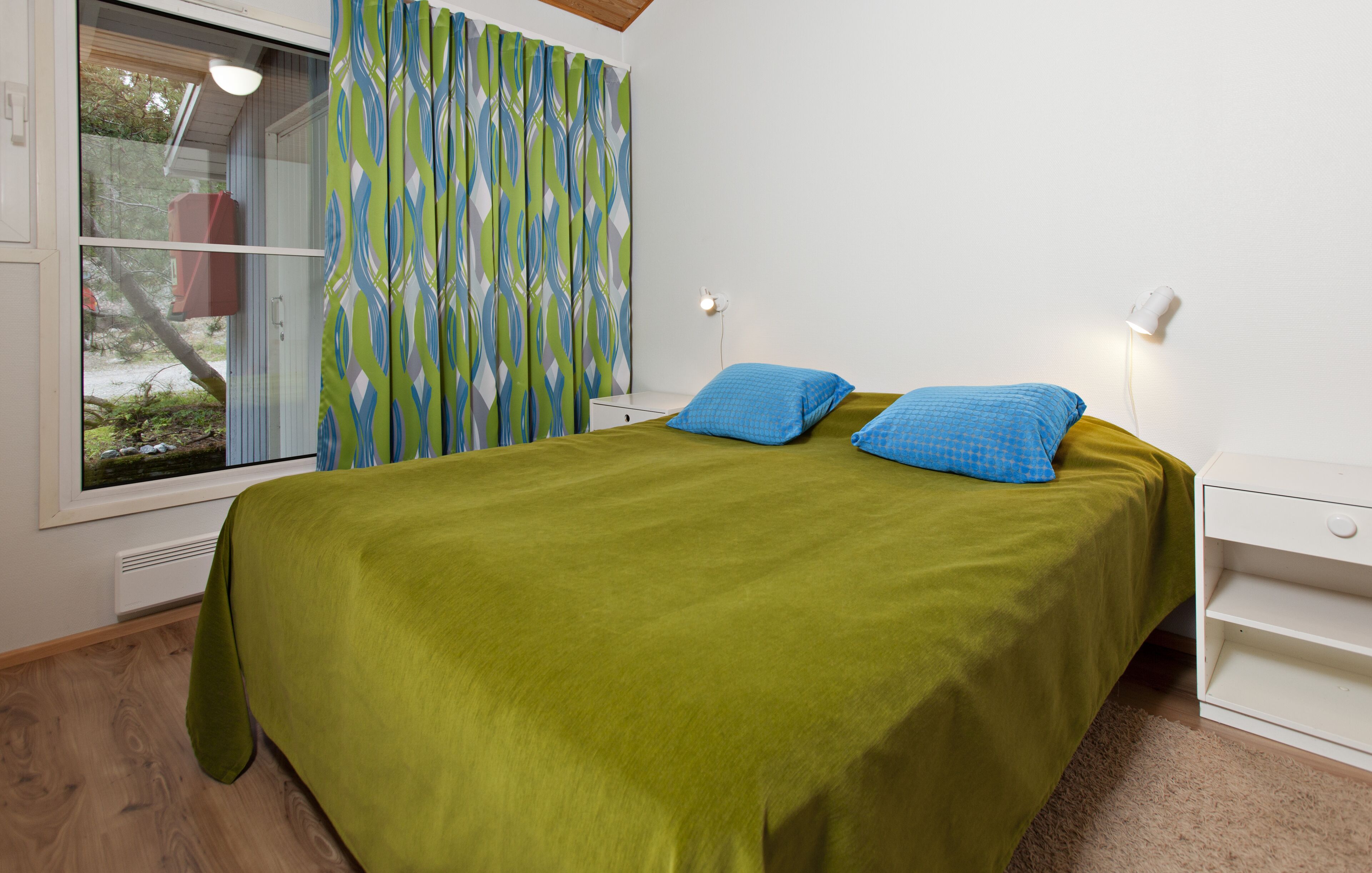 A bedroom with a bed, green bedspread, two blue pillows, a white bedside table, a nightstand, a window with curtains, and a wall-mounted light.
