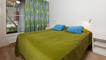 A bedroom with a bed, green bedspread, two blue pillows, a white bedside table, a nightstand, a window with curtains, and a wall-mounted light.