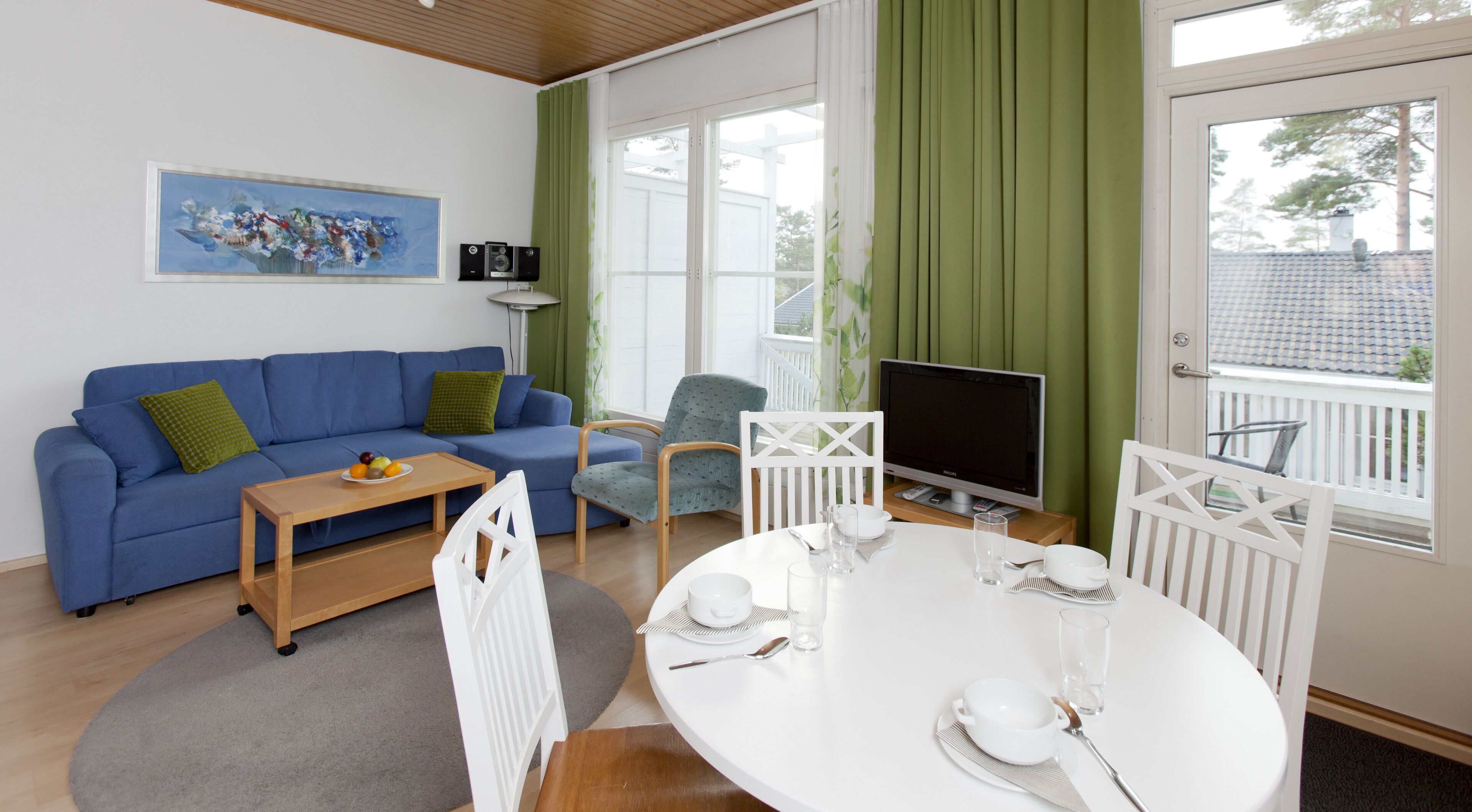 A modern living room with a blue sofa, a dining table set for two, a television, and a window with green curtains.