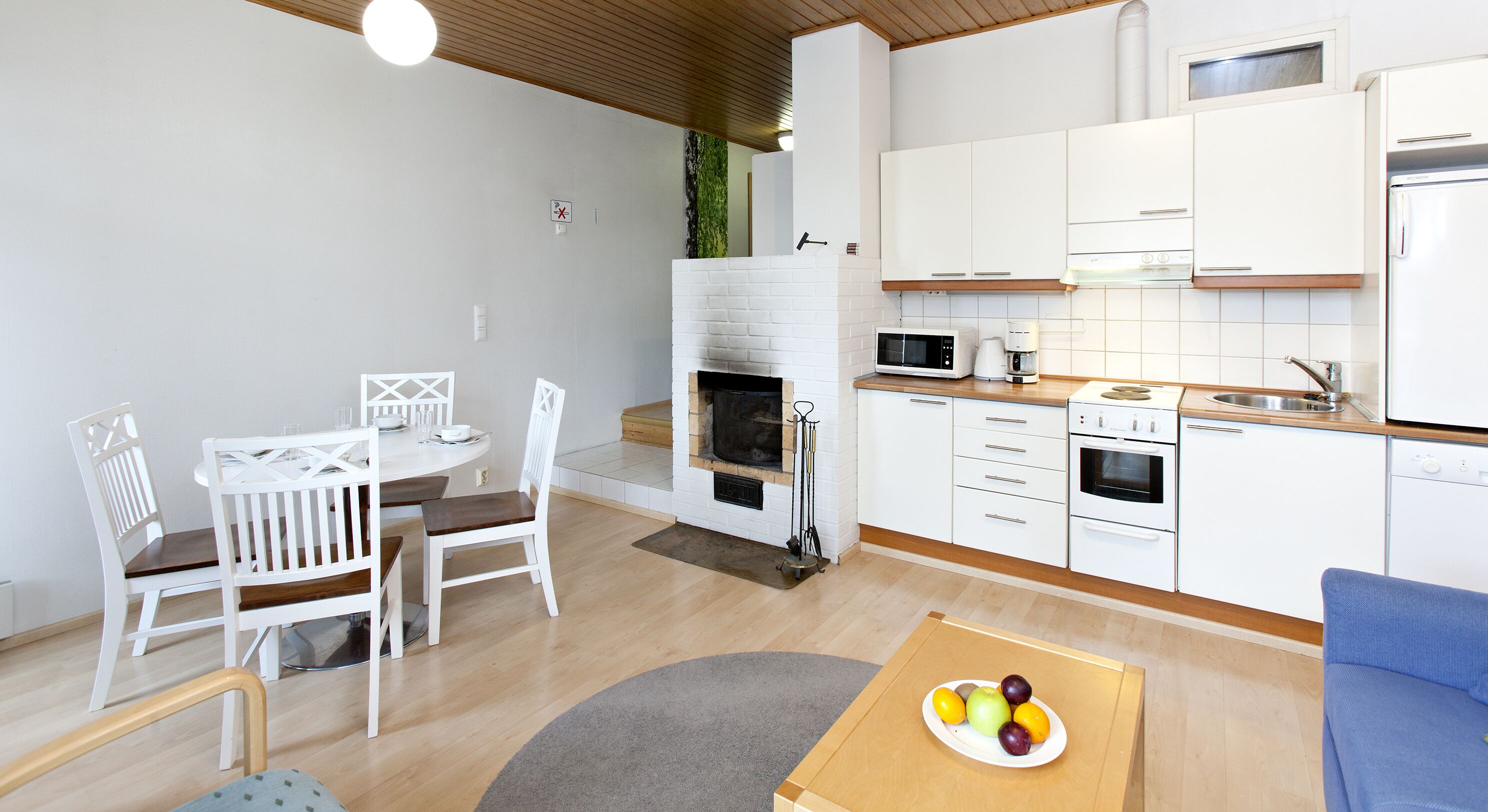 Una cocina moderna con armarios blancos, horno empotrado, microondas y cafetera. Hay un comedor con una mesa y sillas blancas, y un frutero sobre una mesita.