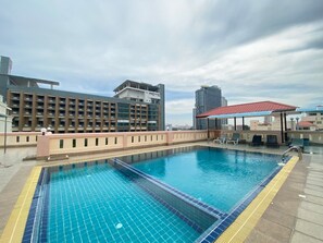 Une piscine sur le toit, avec une terrasse carrelée, un espace salon couvert et des bâtiments modernes en arrière-plan.