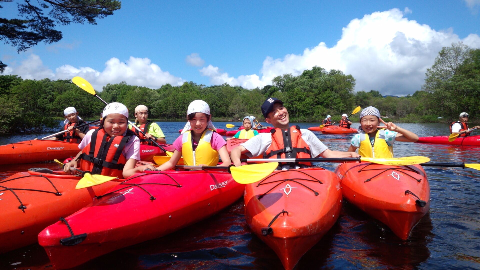 A group of people in red kayaks on a calm body of water, surrounded by trees and a clear sky.