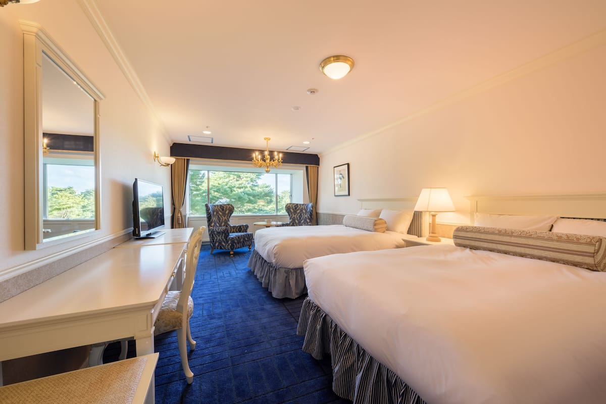A hotel room with two beds, a desk, a chair, a TV, and a window with a view of greenery.