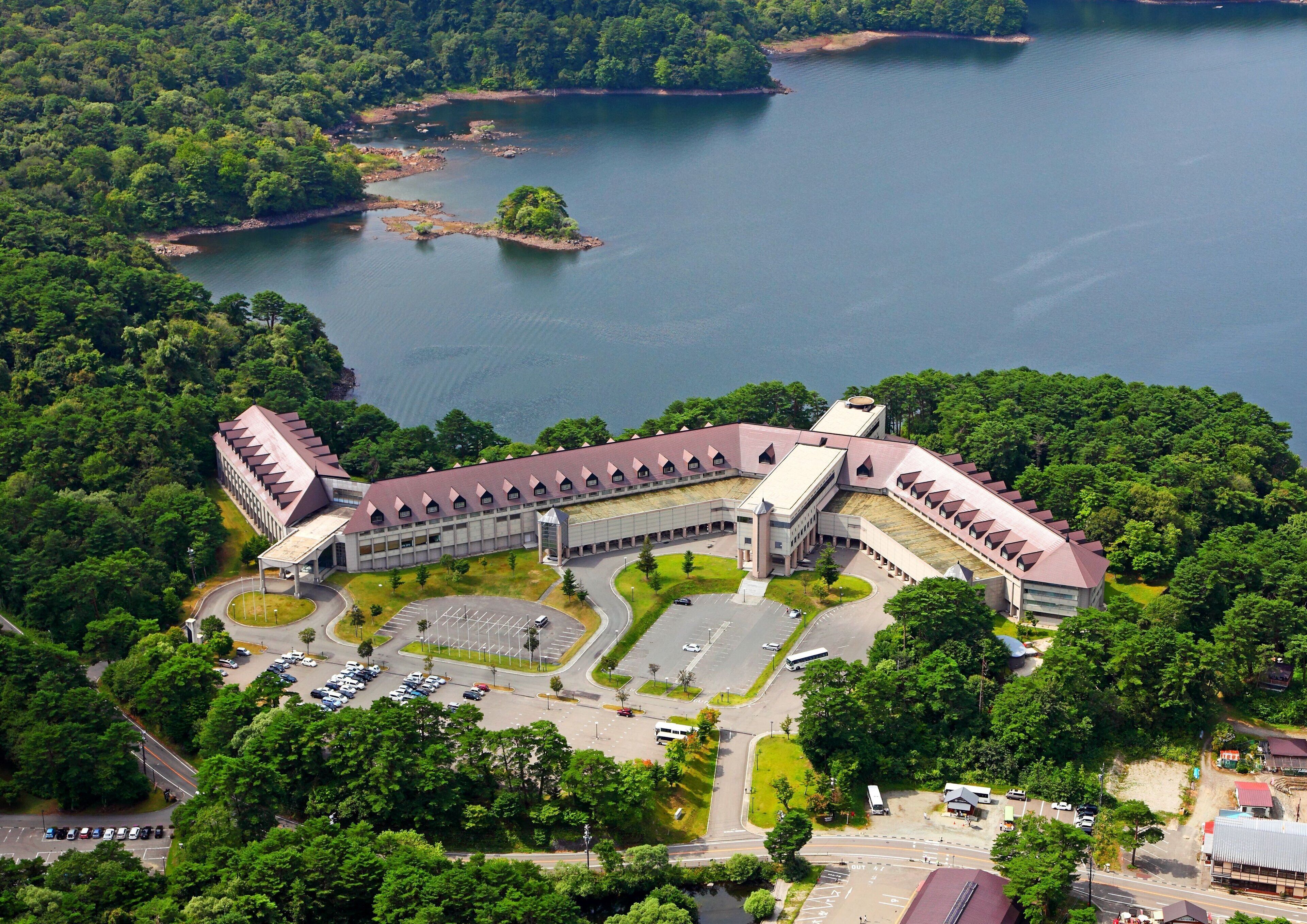 Un grande edificio a più piani con un cortile centrale, circondato da vegetazione rigogliosa e un lago.