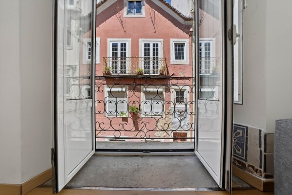 A view through an open door to a balcony with a wrought iron railing, a potted plant, and a building with multiple windows and balconies in the background.
