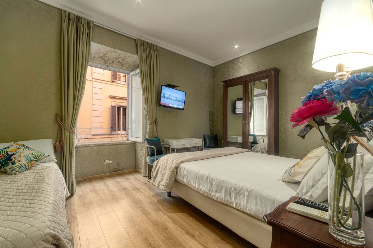 A hotel room with two beds, a desk, a TV, and a vase with flowers.
