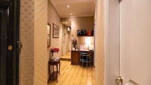 A hallway with patterned wallpaper, a wooden floor, a desk with a chair, and a reception area with a lamp and framed pictures. - Bq House Trevi Luxury Rooms (Rome)