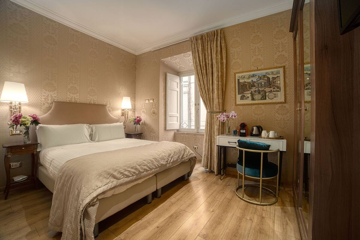 A hotel room with a bed, bedside lamps, a desk with a chair, and a window with curtains.