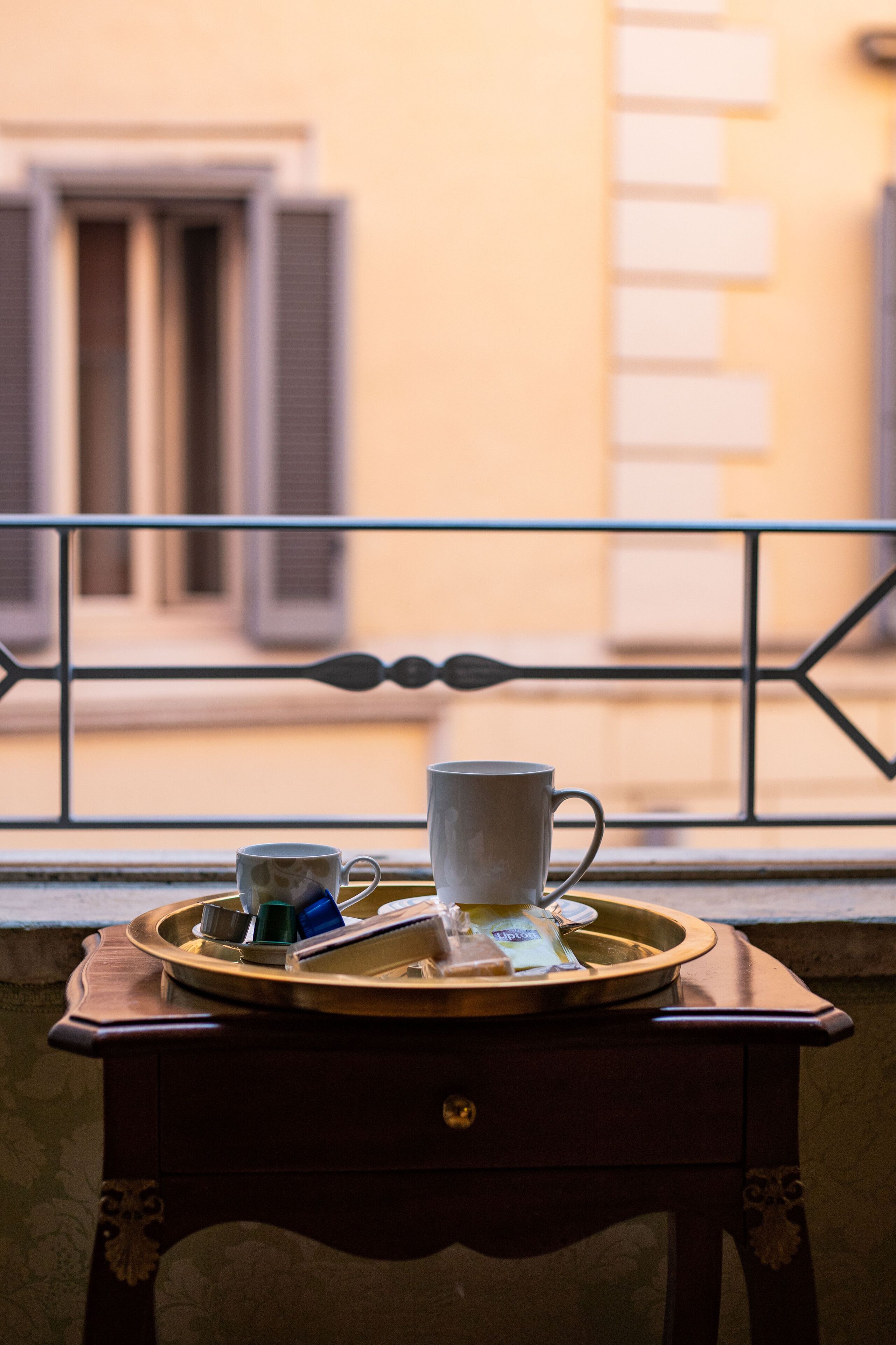 A tray with cups and a teapot on a small table by a window with a balcony railing.