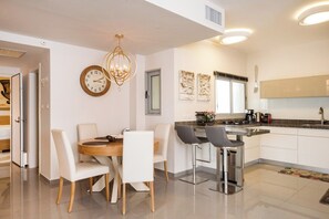 A modern kitchen with a dining area, featuring a chandelier, artwork, and a large window.