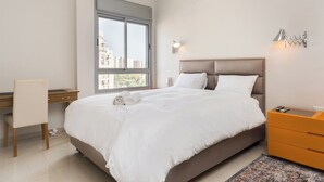 Une chambre à coucher comprenant un lit, un bureau, une chaise, une table de chevet et une fenêtre donnant sur des immeubles.