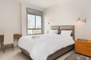 A bedroom with a bed, a desk, a chair, a nightstand, and a window with a view of buildings.