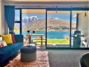 A modern living room with a sofa, ottoman, and a view of a lake and mountains through large glass doors. - Queenstown's Modern Lakefront Marvel - Panoramic BBQ & Unlimited WIFI (Queenstown)