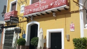 Un edificio amarillo con un letrero que dice 'Posada de Custaquio' y 'Restaurante El Canario'. Hay dos plantas en macetas, una silla blanca y una mesa afuera.