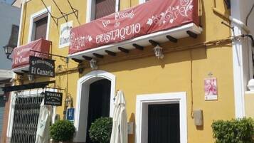 A yellow building with a sign for 'Posada de Custaquio' and 'Restaurante El Canario'. There are two potted plants, a white chair, and a table outside.