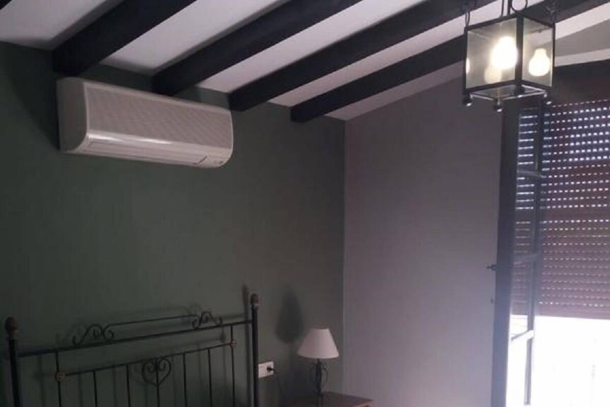 A bedroom with a bed, a nightstand, a lamp, a window with blinds, and an air conditioning unit.