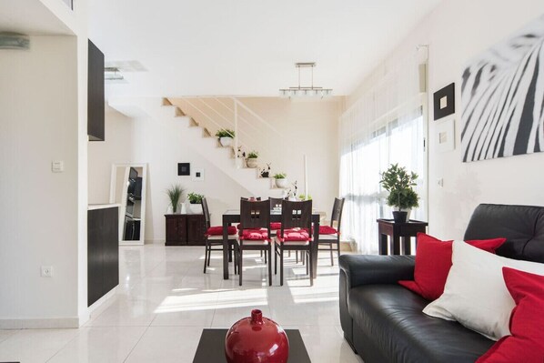 Ein modernes Wohnzimmer mit einem schwarzen Ledersofa, roten und weißen Kissen, einer Essecke mit Tisch und Stühlen sowie einer Treppe mit Blumentöpfen.