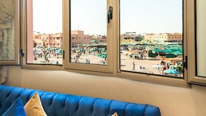 A room with a blue sofa and beige curtains, a view of a bustling outdoor market through large windows. - BNB Medina (Marrakech)