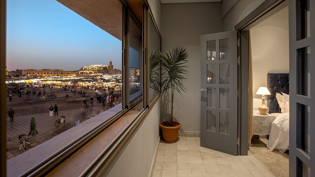 A balcony with a view of a busy square and a building with lights. There is a potted plant and a bed with a lamp. - BNB Medina (Marrakech)