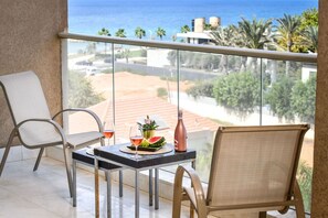 Un balcón con dos sillas, una mesa con una botella y un vaso, y vistas al mar y edificios.