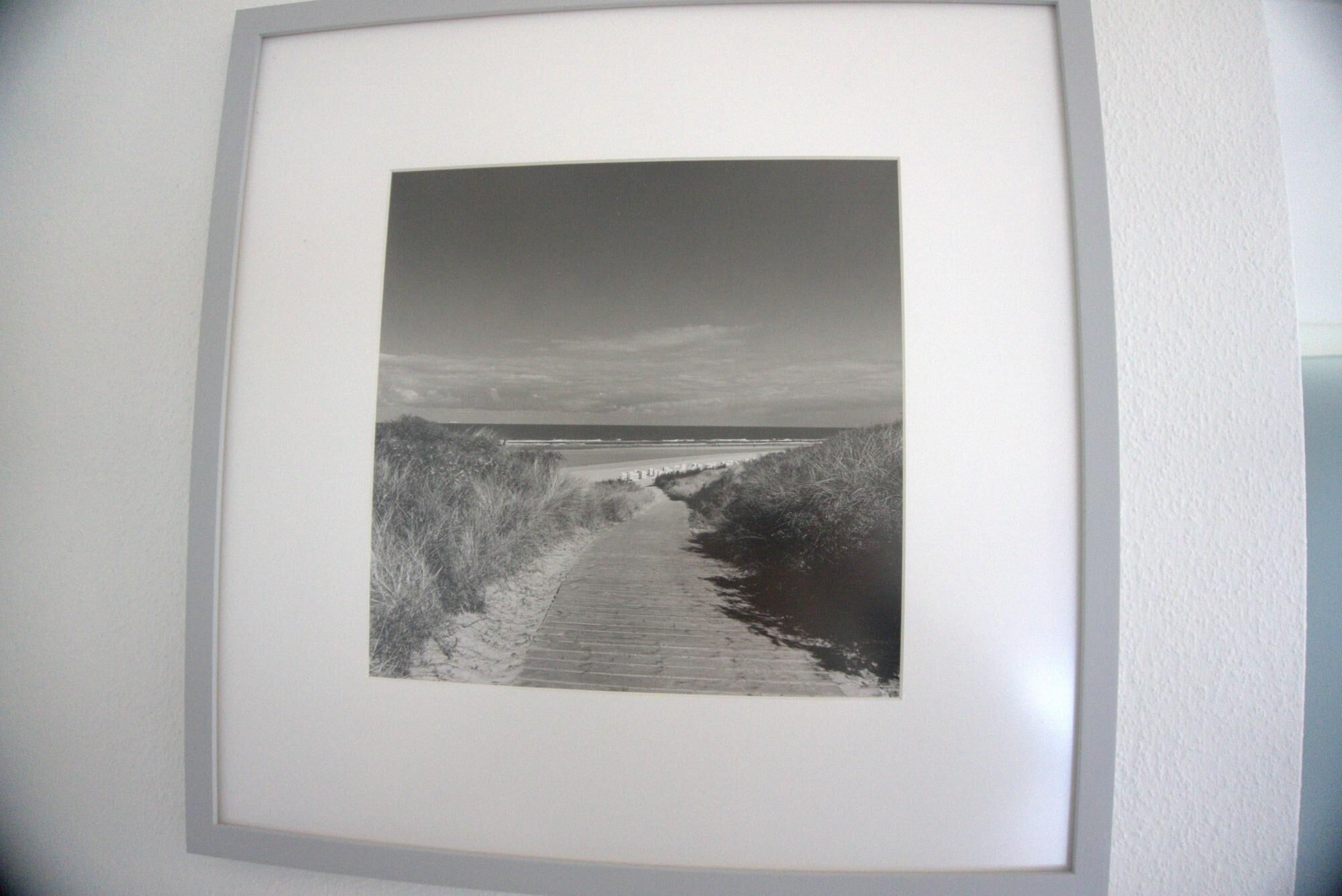 Fotografía en blanco y negro enmarcada, mostrando un sendero costero con vegetación a ambos lados y una vista distante del mar.