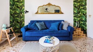 Una habitación con un sofá azul, una mesa de centro redonda blanca, un espejo y una pared con un diseño de hojas.